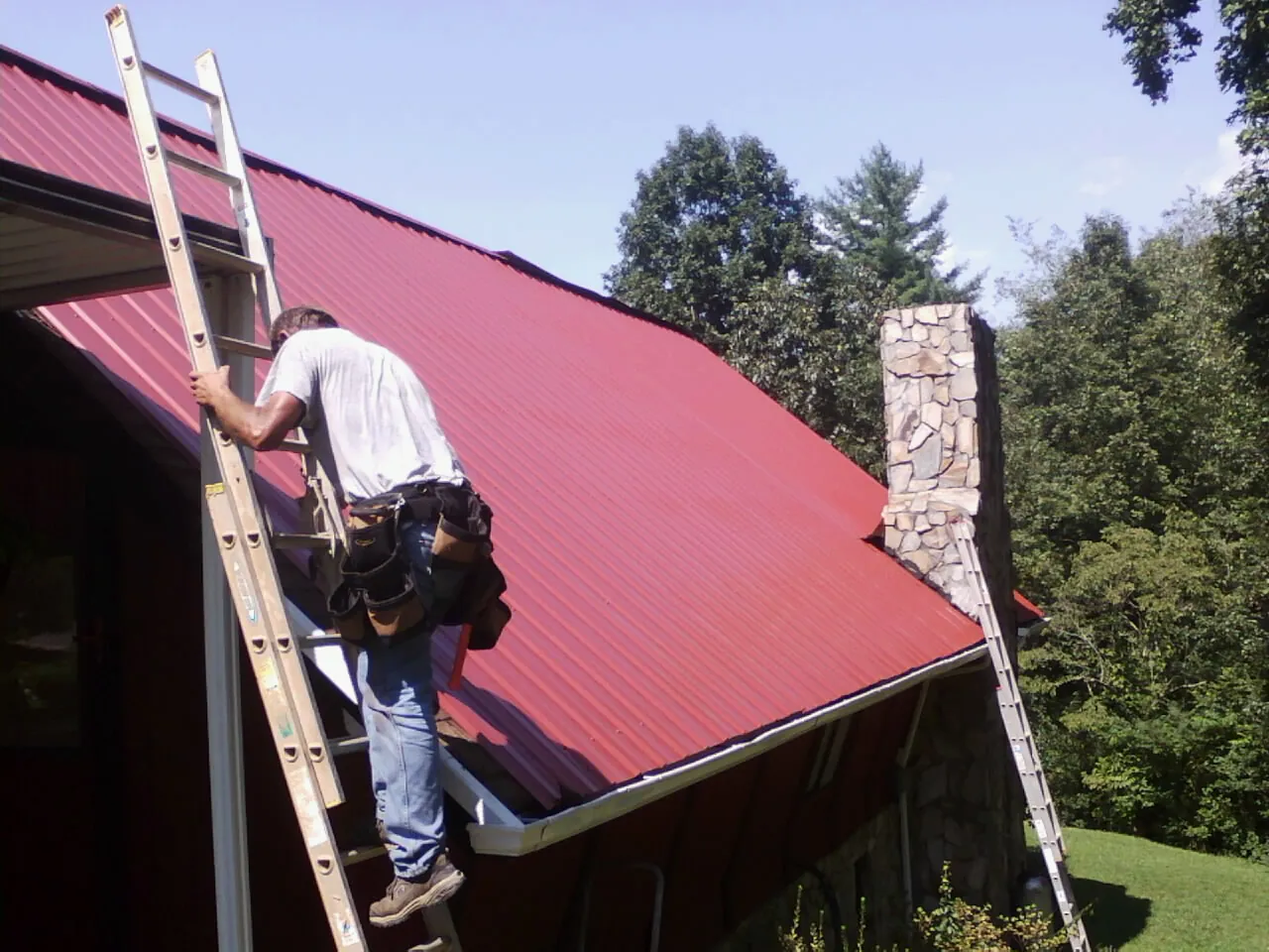 Roofer on a ladder beside a red metal roof and stone chimney — Soffit & Fascia Repair by Roofing Brothers in Park Forest